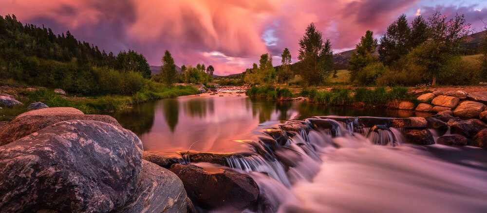 Glassy stream with waterfall foreground, green banks, and sunset rain clouds backdrop.